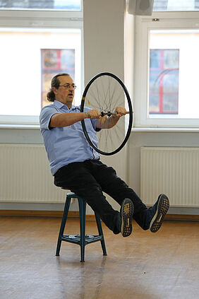 Prof. Böhm sitzt auf einem Drehhocker und hält dabei ein rotierendes Fahrradvorderrad zwischen beiden Händen.