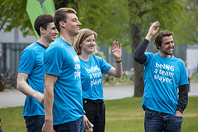 Eine Studentin und drei Studenten tragen türkisfarbene T-Schirts ,it dem Aufdruck "beING a team player." Sie winken Personen außerhalb des Bildes zu.