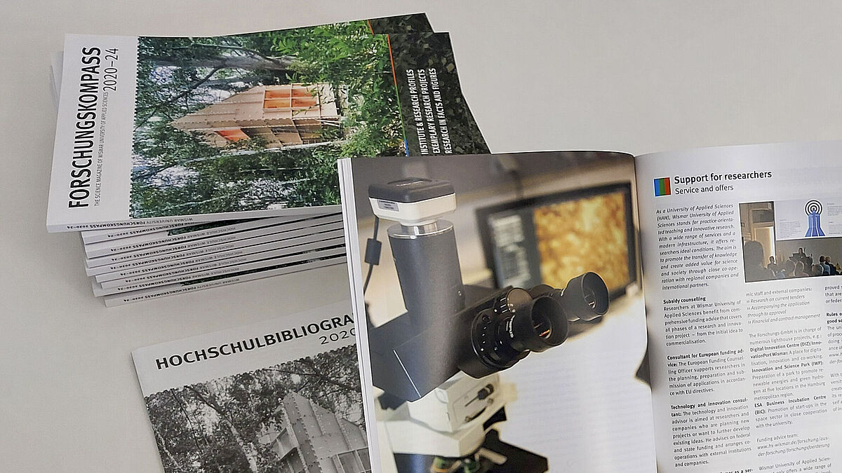 Man schaut einer Person über die Schulter, die eine aufgeblätterte Broschüre in den Händen hält. Auf dem Tisch liegen weiter Exemplare sowie ein Einleger , der etwas kleiner als die im A4-Format gedruckte Broschüre ist.