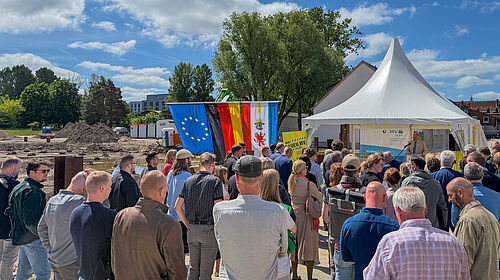 Viele Menschen stehen vor einem Zeltpavillon, mit dem Rücken zur Kamera. Im Zelt steht der Rektor an einem Mikrofon. Links vom Zelt ist die Europafahne, die Deutschlandfahne und die Fahne M-Vs zu sehen. Noch weiter links ist die Baustelle zu erkennen.