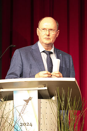 Der Senatsvorsitzende steht auf der Bühne hinter einem Pult, so dass sein Oberkörper zu sehen ist. Er schaut in die Richtung, aus der der Preisträger kommen wird. 