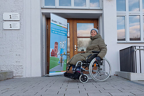 Die KinderUni-Dozentin ist in einem Rollstuhl sitzend an einer Hauseingangstür neben der ein KinderUni-Banner steht, zu sehen.