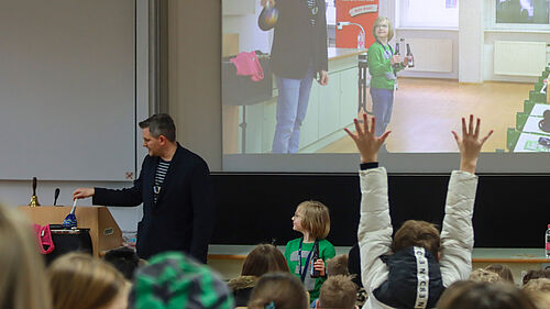 Ein Erwachsener, der KinderUni-Dozent, und ein Kind stehen vor einer Gruppe von Kindern in einem Hörsaal. Der Erwachsene hält einen Gegenstand, zwei Kinder heben die Hände, im Hintergrund ist eine Projektion mit dem gleichen Erwachsenen und Kind zu sehen.
