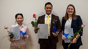 Die drei Personen stehen nebeneinander. Sie halten ihre Urkunde, den Preis und eine Rose in der Hand.