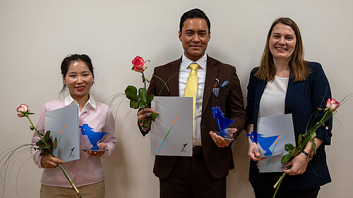 Die drei Personen stehen nebeneinander. Sie halten ihre Urkunde, den Preis und eine Rose in der Hand.