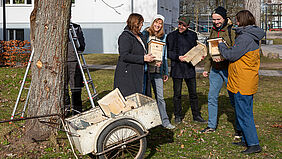 Sechs Personen stehen hinter einer Schubkarre, in der Nistkästen aus Holz liegen. Die Personen halten drei unterschiedliche Nistkästen in der Hand.