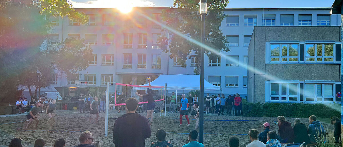 Im Vordergrund ist das Publikum auf dem Rasen und in Liegestühlen sitzend von hinten zu sehen. Sie verfolgen ein Spiel auf dem Volleyballfeld vor ihnen. 