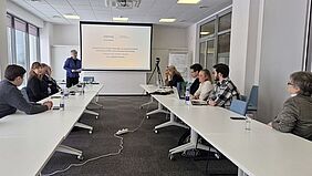 AIKnowIT-Finalkonferenz in Klaipėda Mehrere Projektbeteiligte sitzen an einem U-förmig angeordneten Tisch in einem modernen Seminarraum. Eine Referentin steht vor einer Leinwand mit einer Präsentation zum Interreg-Projekt AIKnowIT. Die Teilnehmenden verfolgen aufmerksam den Vortrag, einige arbeiten mit Laptops.