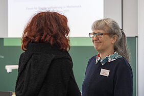 Die Professorin lächelt beim Sprechen. Sie steht einer Person gegenüber, die vonhinten zu sehen ist.