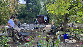 Mehrere Personen arbeiten in einem Garten. Eine Person schiebt eine Schubkarre. Im Hintergrund sind ein Holzhaus sowie Bäume und Büsche zu sehen.