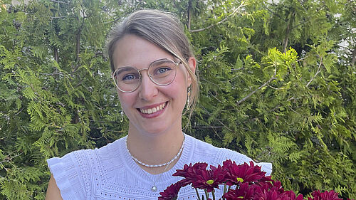 Die Studentin steht vor einer HEcke und hält lächelnd Blumen in der Hand. Sie ist im Porträt zu sehen.