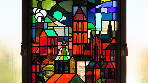 Es ist der Ausschnitt eines farbigen Bleiglasfensters zu sehen, auf dem dem z. B. Dächer und eine Kirche zu erkennen sind.