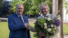 Der Rektor hält einen großen Blumenstrauß in der linken Hand. Mit der rechten Hand nimmt er die Glückwünsche des Senatsvorsitzenden entgegen, der links von ihm steht.
