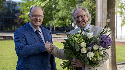 Der Rektor hält einen großen Blumenstrauß in der linken Hand. Mit der rechten Hand nimmt er die Glückwünsche des Senatsvorsitzenden entgegen, der links von ihm steht.