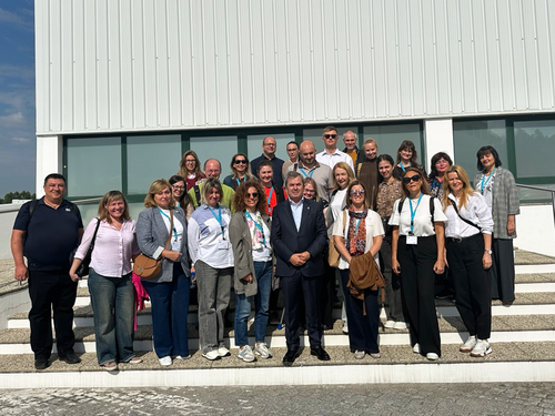 A group of approximately 25 people stands in several rows on the steps of a modern building with a glass façade and white cladding. All participants wear name badges, some with blue lanyards.
