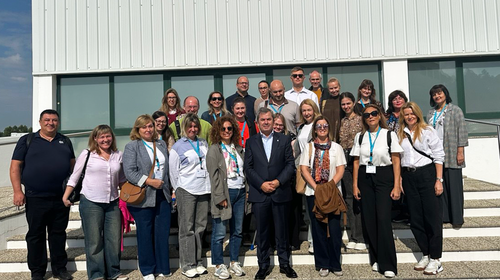 A group of approximately 25 people stands in several rows on the steps of a modern building with a glass façade and white cladding. All participants wear name badges, some with blue lanyards.