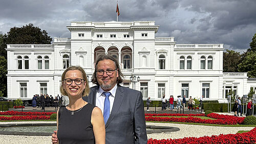 Zwei Personen in formeller Kleidung stehen vor der Villa Hammerschmidt mit Flagge auf dem Dach und einem Garten mit roten Blumen im Vordergrund