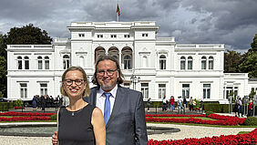 Zwei Personen in formeller Kleidung stehen vor der Villa Hammerschmidt mit Flagge auf dem Dach und einem Garten mit roten Blumen im Vordergrund