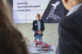 Der Fotograf schaut über die Schulter zweier Stipendiaten. Der Prorektor steht vor einer Bildschirmpräsentation auf der Datum und Veranstaltungstitel stehen.