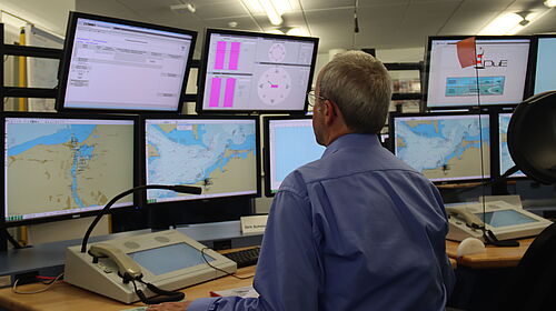 Es sind neune Bildschirme mit Seekarten und Informationen im VTSS-Simulator zu sehen. Vor diesen sitzt ein Mann, der von hinten zu sehen ist.