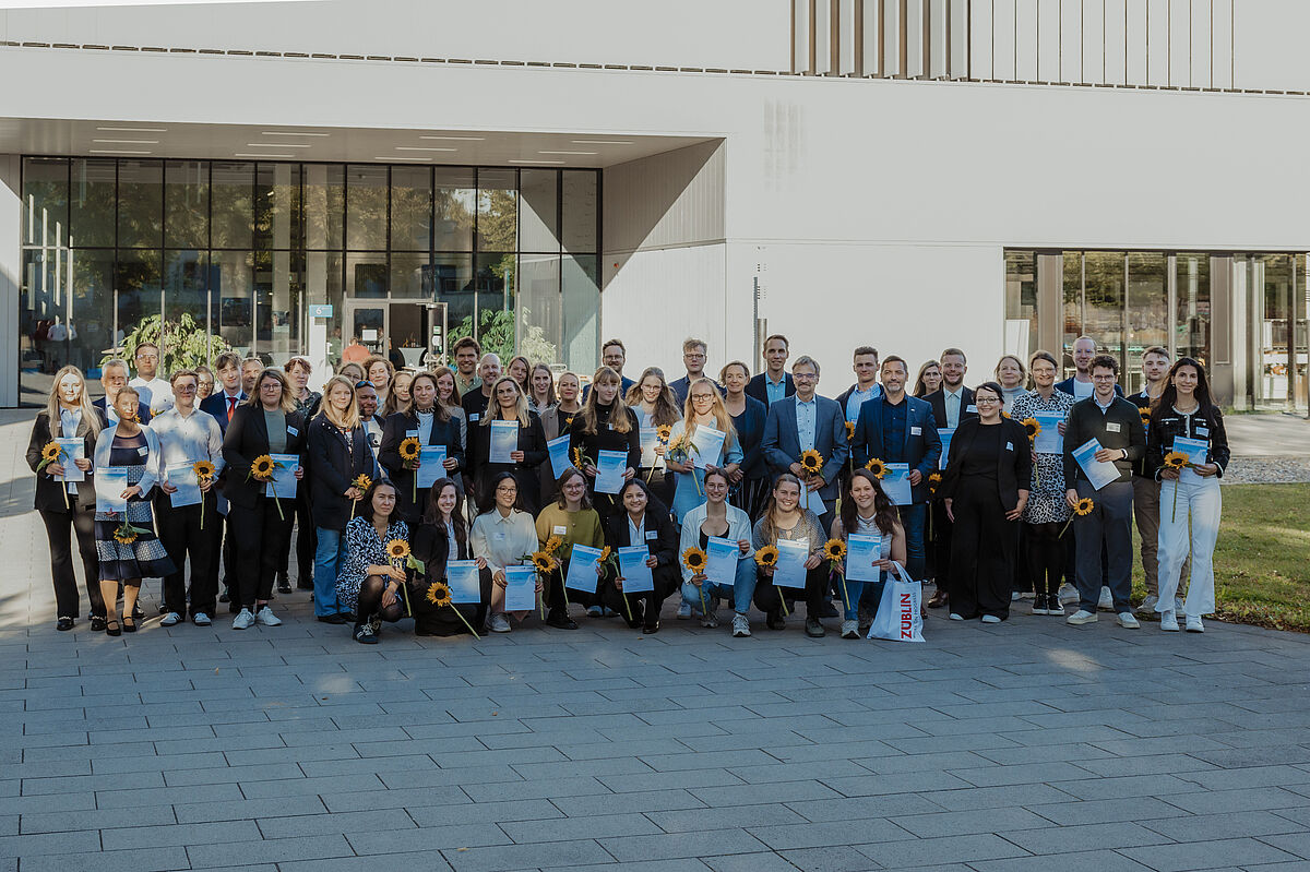 50 Personen stehen neben- und hintereinander. Einige halten Blumen und Urkunden in den Händen. Daran erkennt man die Studierenden. Im Hintergrund ist Haus 6 zu erkennen.