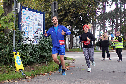 Eine Läuferin und ein Läufer sind auf dem Weg in die Gartenanlage und zeigen lächelnd "DAumen hoch".