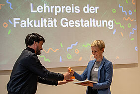 Die beiden PErsonen stehen sich gegenüber. Sie sind von der Seite zu sehen, wie sie gerade beide die Preisskulptur in der Hand halten.