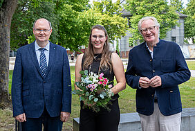 Drei Personen stehen auf dem Wismarer Campus, auf dem im Hintergrund Bäume mit frischem Grün zu sehen sind. Die mittlere Person ist die gewählte Prorektorin. Sie hält einen Blumenstrauß, alle drei tragen formelle Kleidung.
