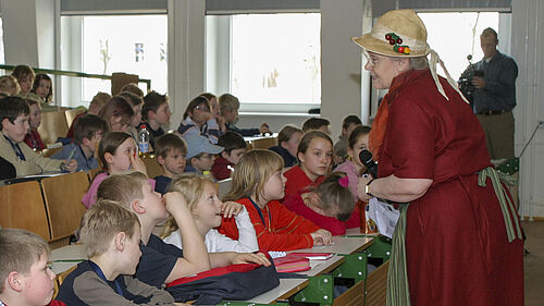 Die Dozentin steht mit einem Mikrofon in der Hand und mit einem roten Kleid sowie Strohhut bekleidet vor der ersten Reihe und lächelt ein Kind in der zweiten Reihe an.