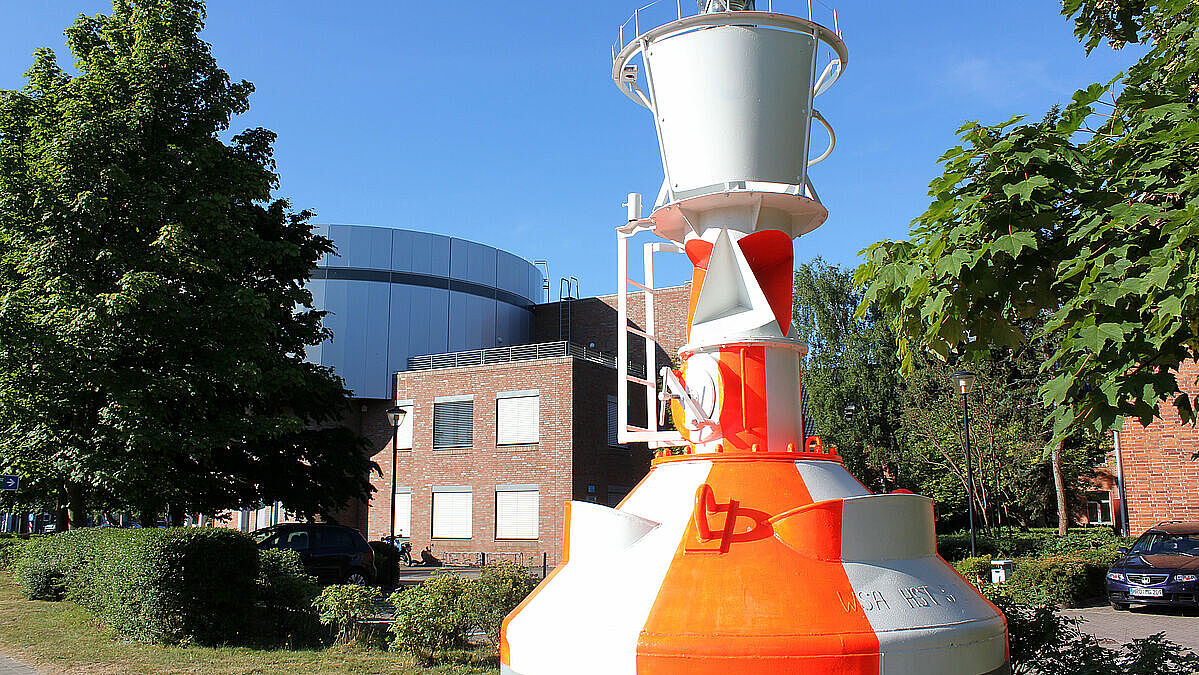 Ein weiß-orange lakiertes Seezeichen steht auf einem Platz, im Hintergrund mehrere Gebäude und Bäume