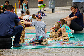 Im Vordergrund sind Kinder zu sehen, die mit Holzklötzen Türme bauen. Erwachsene sitzen entspannt daneben. Im Hintergrund sieht man unscharf weitere Kinder spielen.