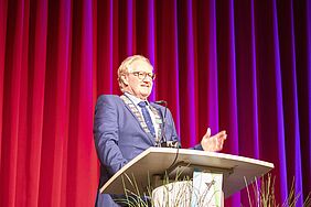Der Rektor steht auf der Bühne hinter einem Pult, so dass sein Oberkörper zu sehen ist. Er trägt die Rektorkette.