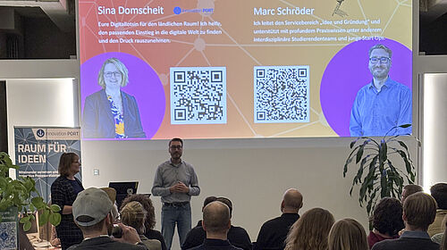 Man schaut über die Köpfe von 13 PErsonen hinweg auf eine Präsentation. Davor stehen Sina Domscheit und Marc Schröder.