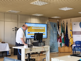 A presenter stands in front of a screen displaying the interactive coursebook on “Compliance Management.” Several flags and project banners are visible in the background.