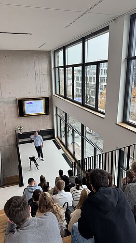 Lecture on brand management at Paulihaus A group of students sit on wooden steps and follow a lecture in front of a screen in an open room with floor-to-ceiling windows.