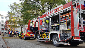 Es ist ein großes Löschfahrzeug zu sehen, dessen drei Seitenrollos geöffnet sind. Vor dem steht ein kleines Transportfahrzeug der Feuerwehr. ImHintergrund ist das Haus 1 zu sehen.