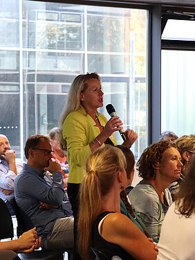 Eine Frau steht mitten im Publikum, das auf Stühlen sitzt. Sie hält ein Mikrofon in der Hand.