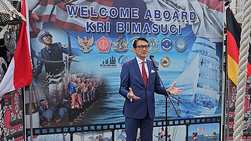 Der Botschafter steht vor einem typisch indonesischem Banner, auf dem "Welcome Aboard Kri Bima Suci" steht, und soricht ins Mikrofon.