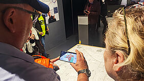 Über die Schulter eines Mannes hinweg sieht man das Display eines Handes. Darauf ist ein 3D-Modell zu erkennen, auf dem Tisch darunter eine Grundrisszeichnung.