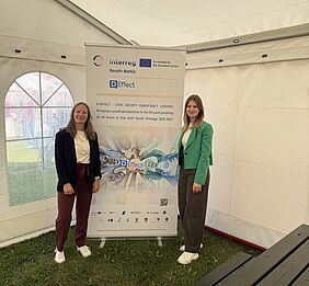 Hochschule Wismar Representatives at the D-EFFECT Festival. Two women stand inside a white event tent in front of a project banner displaying information about the D-EFFECT project.