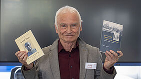 Der Professor schaut in die Kamera und hält in jeder Hand ein Buch hoch.