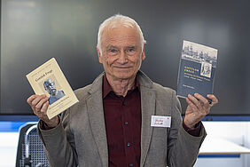 Der Professor schaut in die Kamera und hält in jeder Hand ein Buch hoch.
