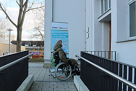 Die KinderUni-Dozentin ist in einem Rollstuhl sitzend an einer Hauseingangstür neben der ein KinderUni-Banner steht, zu sehen.