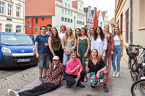 Die Personen stehen als Gruppe auf dem Fußweg. Ein Student liegt schräg aufgestützt und zwei Studentinnen prosten sich zu. Im Hintergrund ist eine typisch hanseatische Häuserzeile zu sehen.