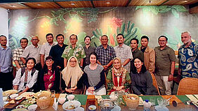 19 Personen stehen bzw. sitzen hinter einem langen Tisch. Auf diesem befinden sich zahlreiche leere Schüsseln, Teller und Gläser.