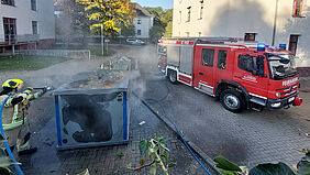 Es ist ein Feuerwehrmann zu sehen, wie er mit einem Wasserstrahl den Brand in der Tonne löscht. Daneben steht das Löschfahrzeug. 