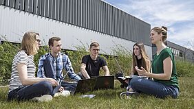 Es sind fünf Studierende mit Laptops zu sehen, die auf dem Rasen vor dem Haus 6 sitzen und miteinander sprechen. 
