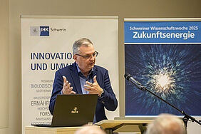 DEr REdner steht vor einem Banner mit der Grafik der Schweriner Wissenschaftswoche 2025 und gestikuliert mit seinen Händen.