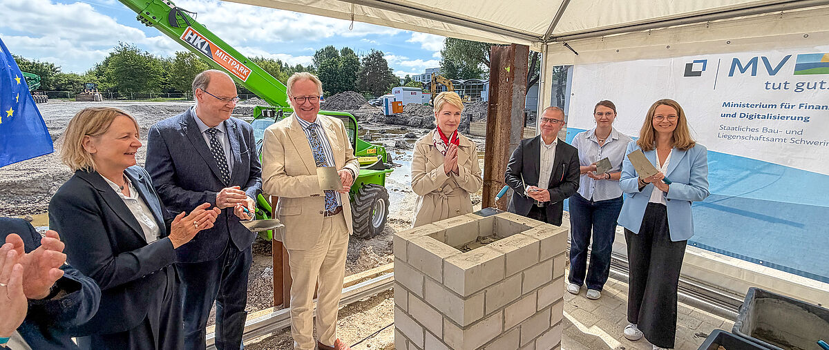 Mehrere offizielle Vertreter*innen am Grundstein eines neuen Gebäudes mit Baukellen in der Hand, im Hintergrund Banner von MV und EU