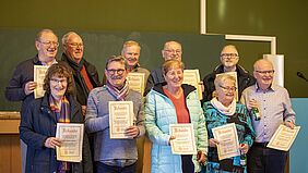 Die Personen stehen in zwei Reihen auf einem Podium und halten ihre neuen und alten Urkunden in die Kamera.